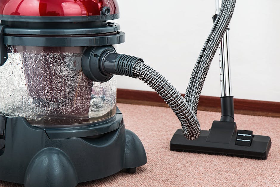Close-up of a cordless vacuum cleaner with a transparent dust container filled with dust and debris, connected to a flexible, patterned hose. The vacuum is positioned on a pinkish-beige carpet in a re